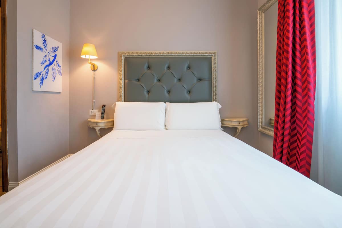 Una habitación de hotel con una cama grande, un cabecero con diseño de rombos, una lámpara de noche, un espejo y una cortina roja.
