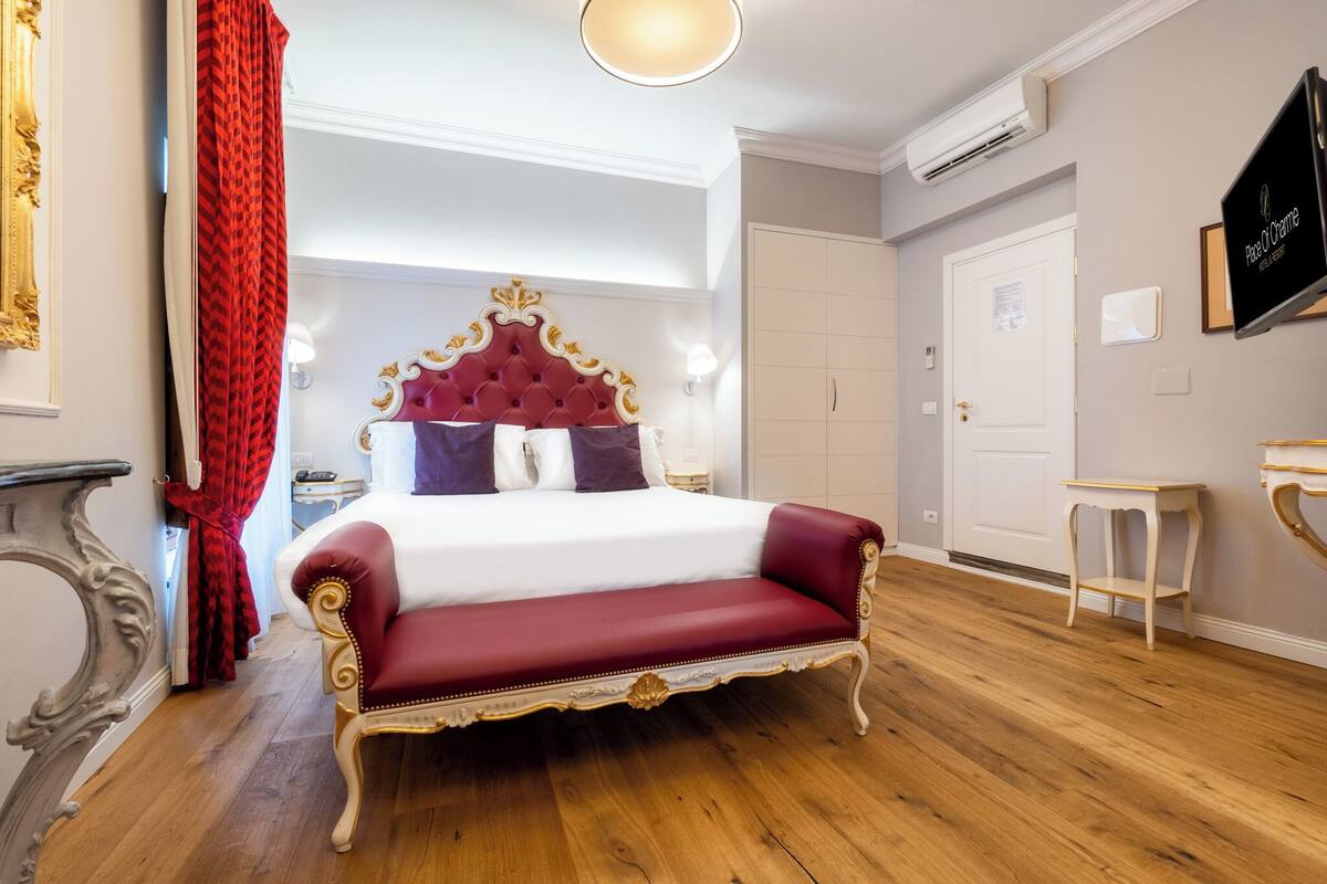 Un dormitorio amplio con una cama grande, un banco tapizado en rojo, un cabecero clásico ornamentado y suelo de madera.