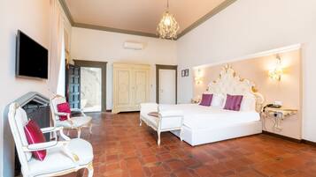 A spacious bedroom with a large bed, two white armchairs, a chandelier, and a fireplace.