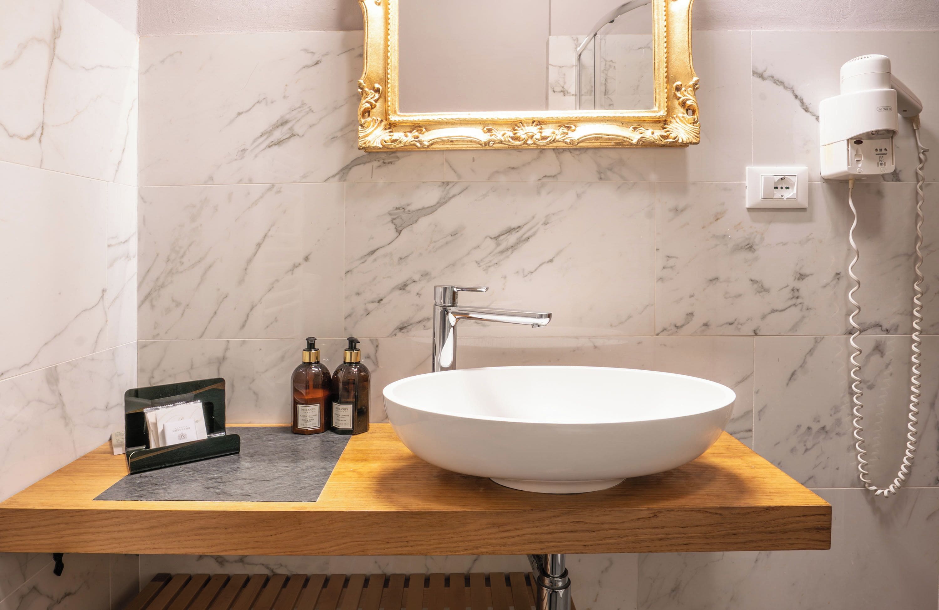Un baño con pared de mármol, tocador de madera, lavamanos blanco, espejo con marco dorado, un secador de pelo y una pequeña botella de champú.