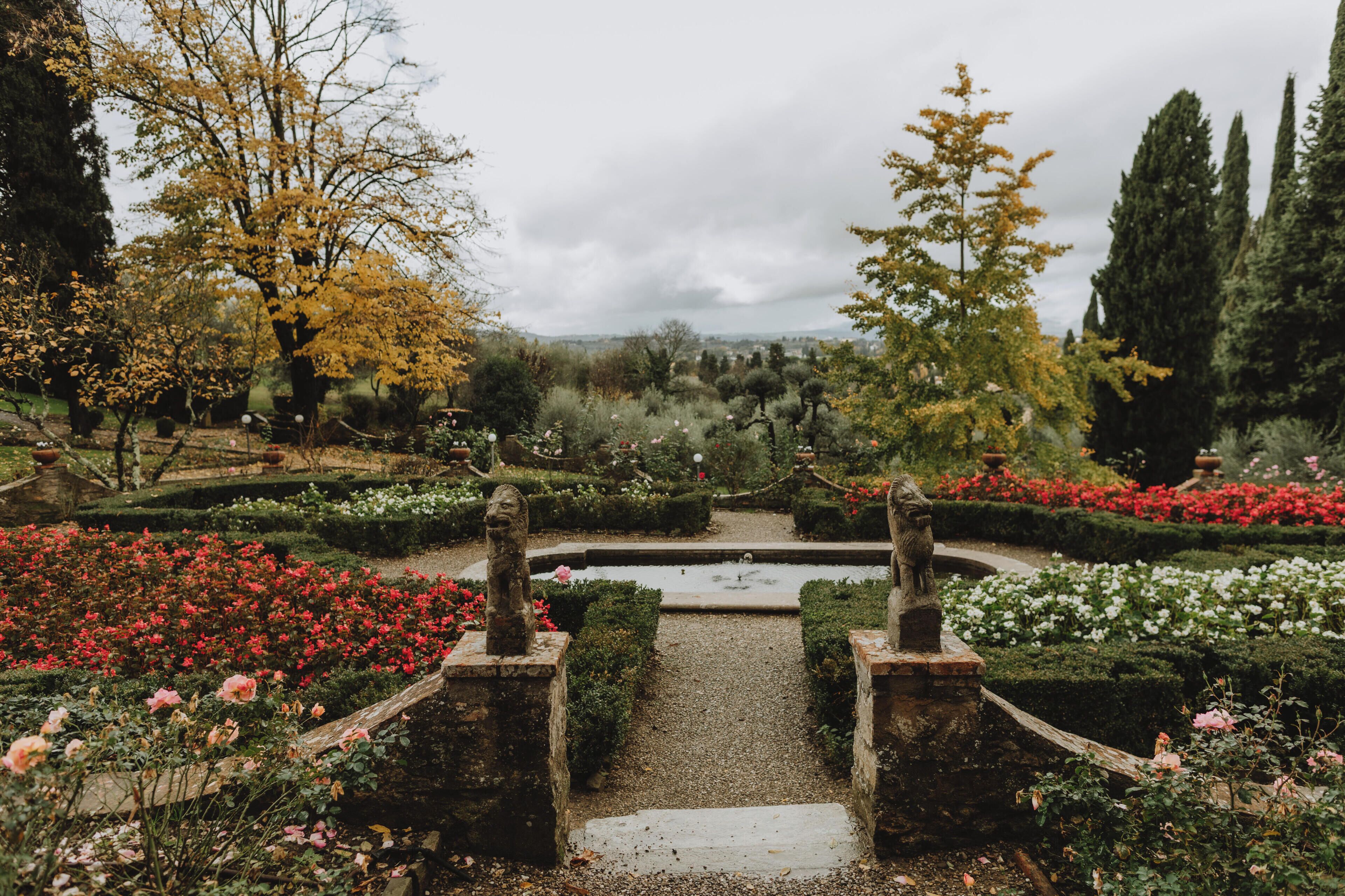 Ein Gartenweg mit Statuen, umgeben von verschiedenen Blumenbeeten und Bäumen.