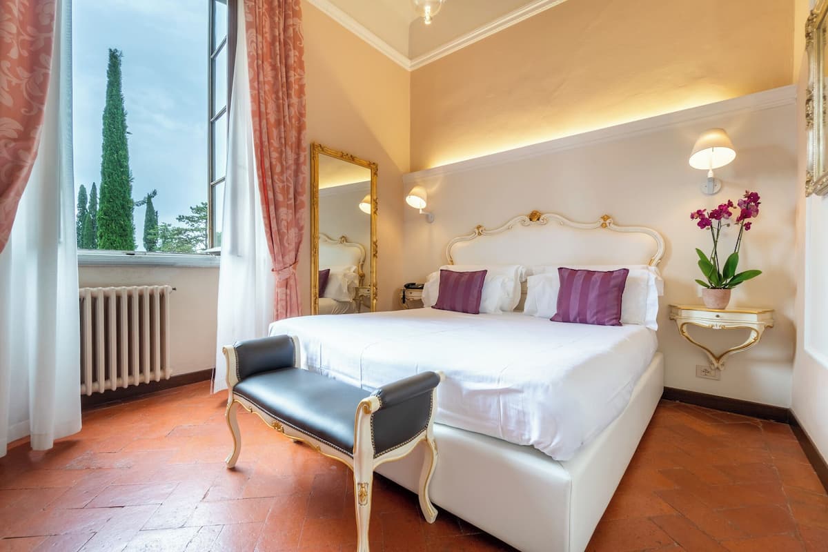 Habitación de hotel con una cama grande, una silla, un espejo, una mesita de noche con lámpara y vistas a árboles.