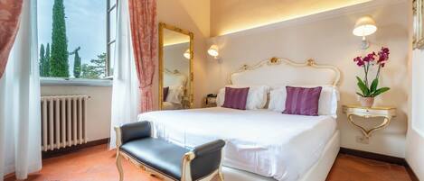 A hotel room with a large bed, a chair, a mirror, a bedside table with a lamp, and a view of trees outside.