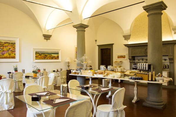 Un comedor con mesas y sillas de vidrio, un sistema de buffet y arquitectura clásica con columnas y arcos.