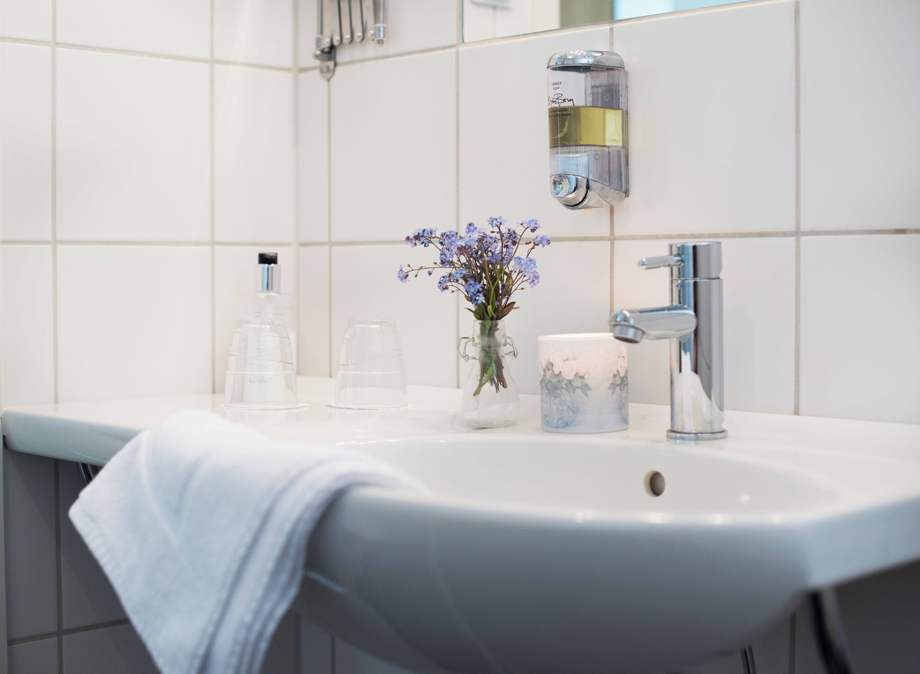 A bathroom with a white sink, a mirror, a soap dispenser, and a vase with flowers.