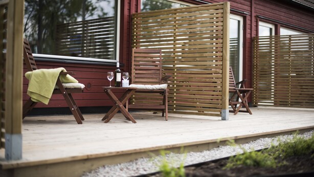 A wooden deck with two chairs and a table, a wooden fence, and a house with large windows. - Toftastrand Hotell (Växjö)