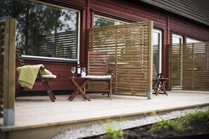 A wooden deck with two chairs and a table, a wooden fence, and a house with large windows. - Toftastrand Hotell (Växjö)