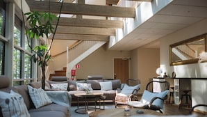 A spacious living area with a sofa, chairs, a coffee table, and a fireplace. There is a staircase leading to an upper level and a large window providing natural light. - Toftastrand Hotell (Växjö)