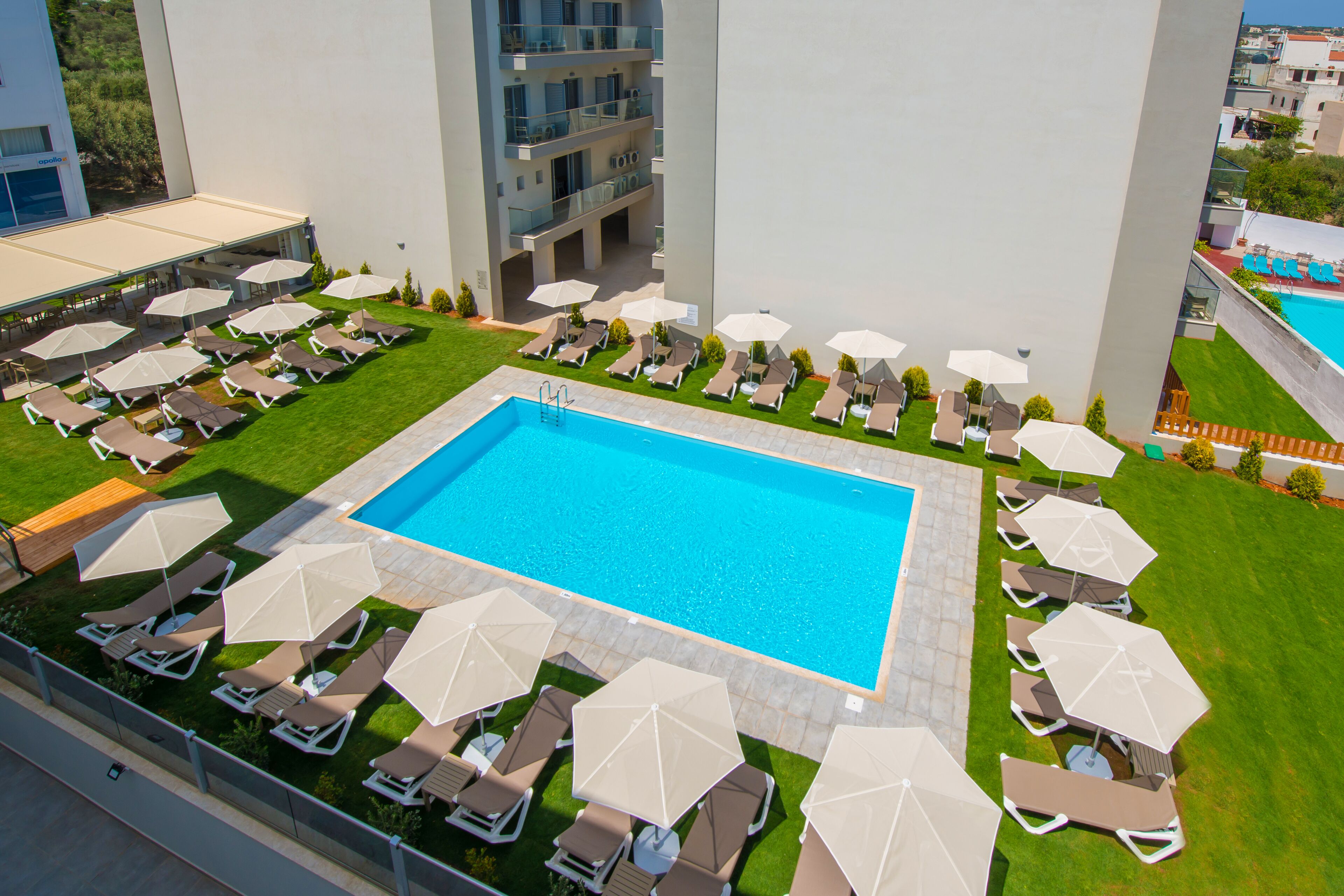 A rooftop pool area with multiple lounge chairs and umbrellas, surrounded by a well-maintained lawn and a building with balconies.