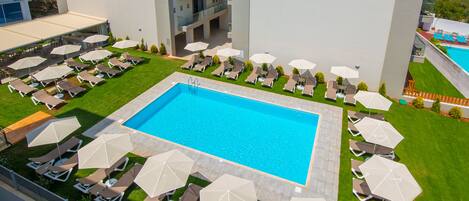 A rooftop pool area with multiple lounge chairs and umbrellas, surrounded by a well-maintained lawn and a building with balconies.