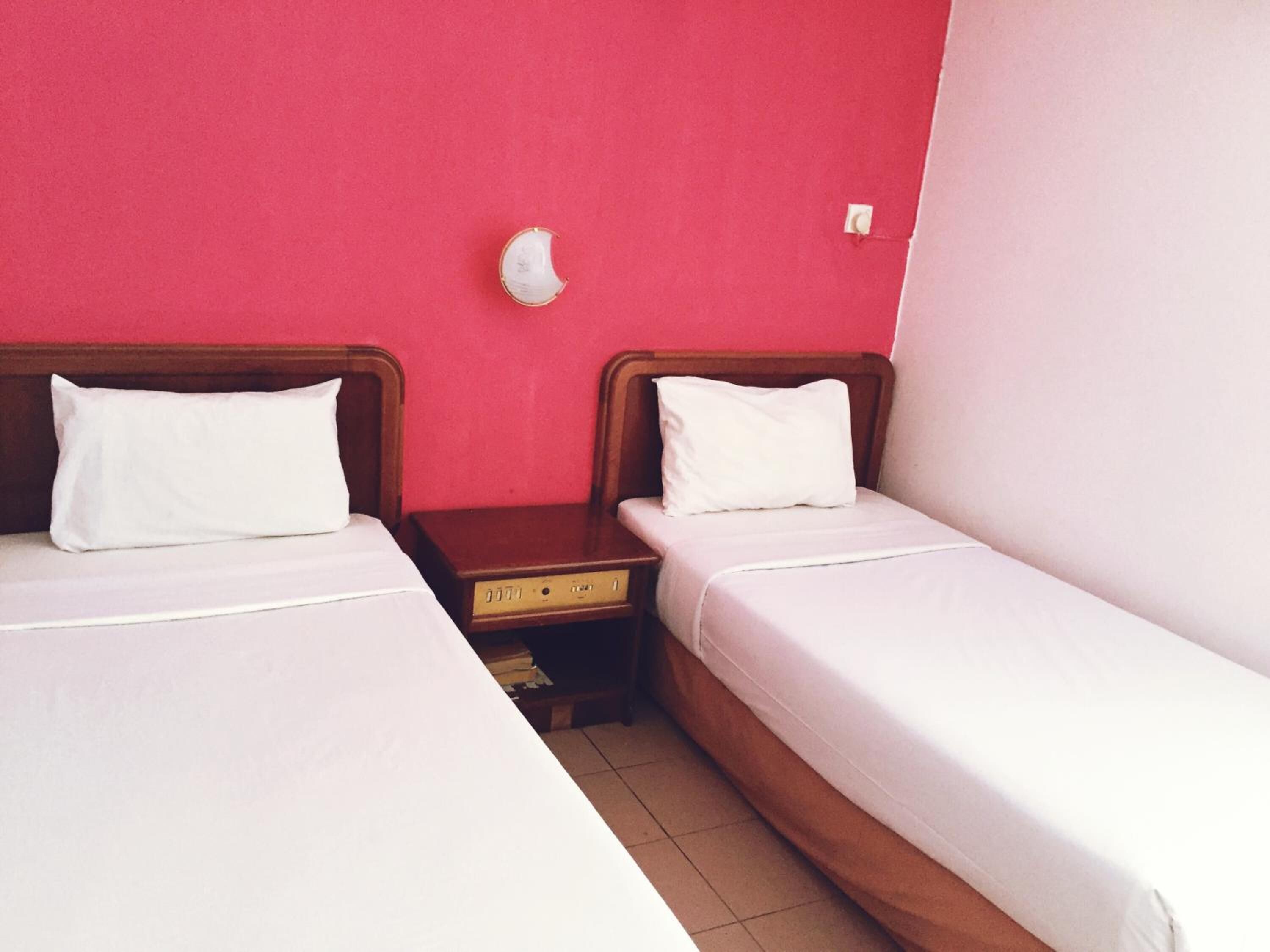 A room with two single beds, wooden headboards, white bedding, a wooden nightstand with a radio, and a pink wall.