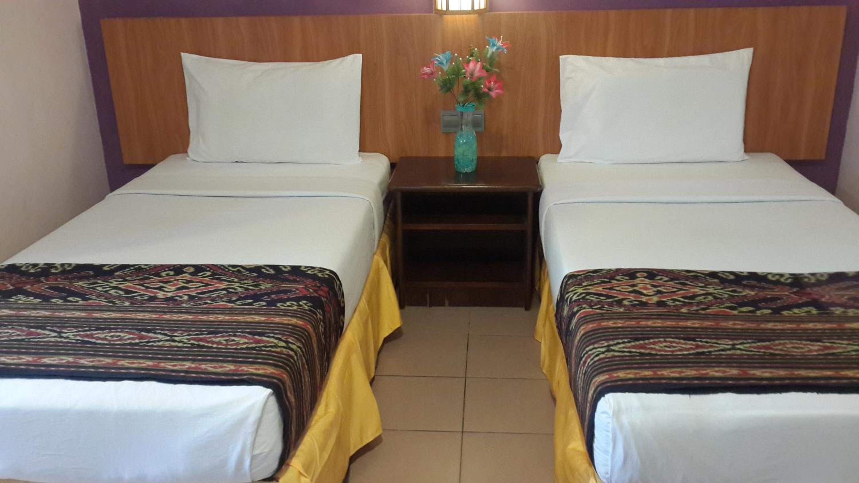A hotel room with two beds, each with a patterned bedspread and white pillows. There is a nightstand with a vase of flowers between the beds.