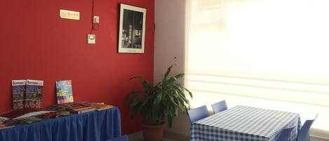 A room with a red wall, a large window, a table with a blue tablecloth, and a potted plant.