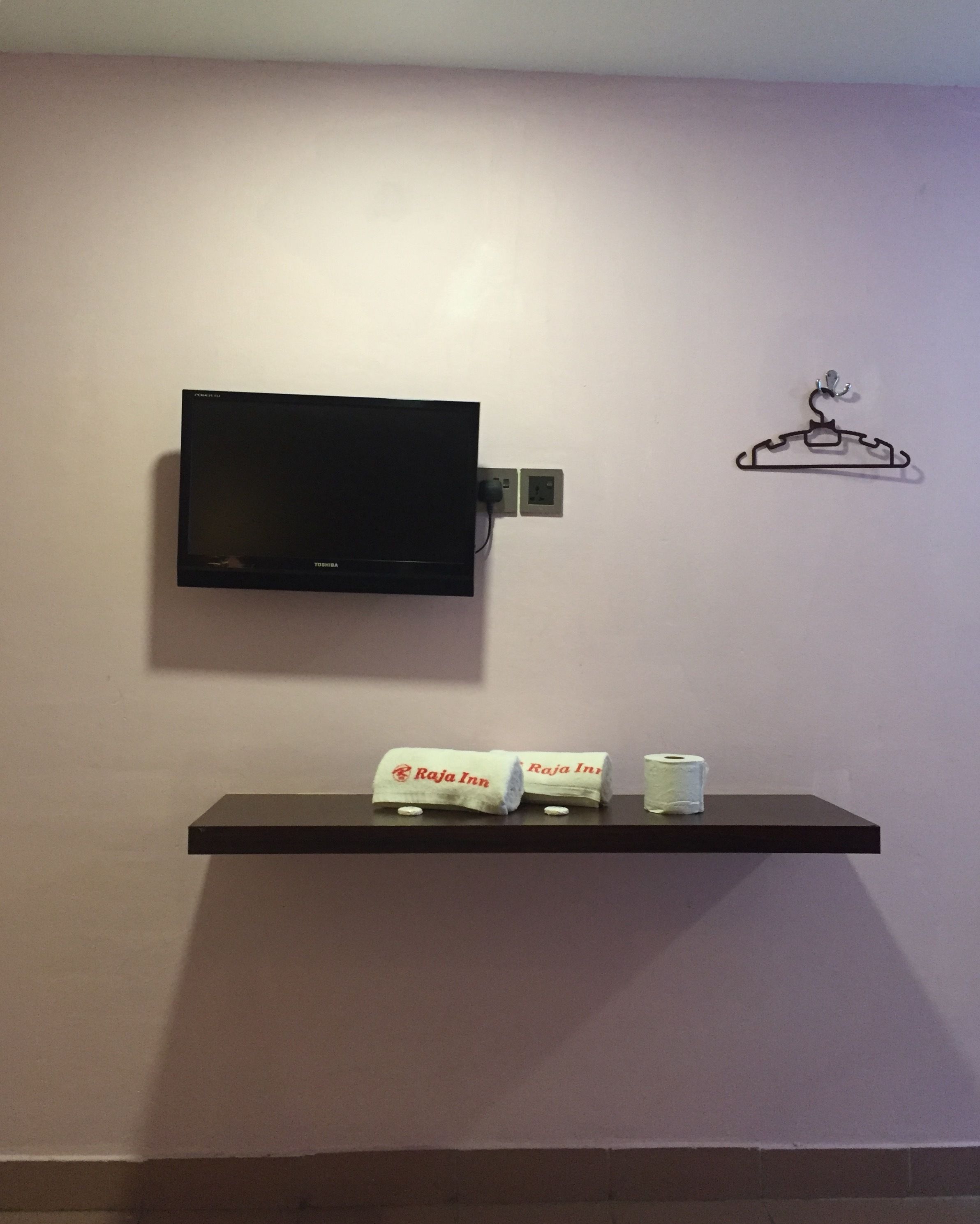 Un téléviseur à écran plat fixé au mur, une étagère noire en dessous, sur laquelle sont posés deux serviettes enroulées et un rouleau de papier toilette.