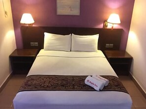 A hotel room with a bed, two bedside lamps, a nightstand, a patterned throw blanket, and a rolled towel with text.