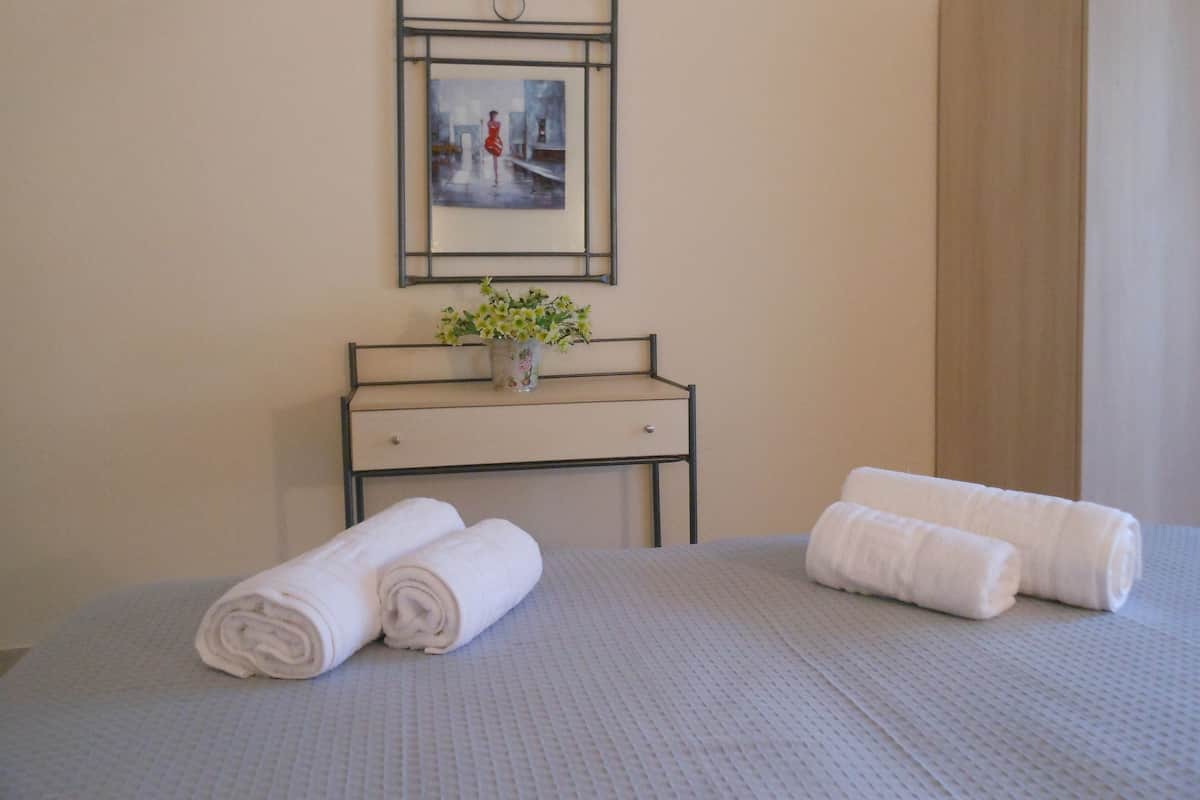 A neatly made bed with rolled white towels, a small bedside table with a vase of flowers, and a framed picture on the wall.