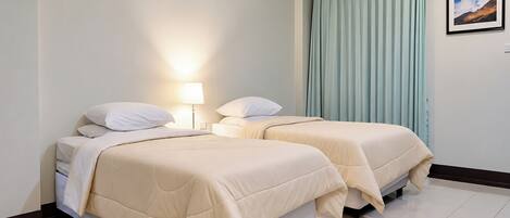 A hotel room with two beds, a nightstand, a lamp, a window with curtains, and a wall-mounted air conditioner.