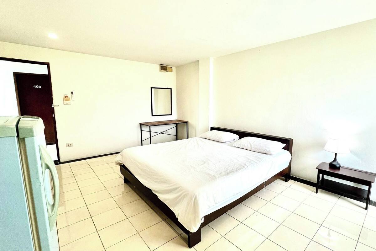 Habitación de hotel con una cama, una mesita de noche con lámpara, un pequeño atril y una puerta con el número 408.