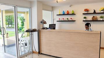 A reception desk with a glass door entrance, a small table with a lamp, and shelves with potted plants and decorative items.