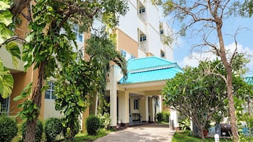 Entrada de un hotel con un paseo techado, rodeada de vegetación y un cielo despejado.