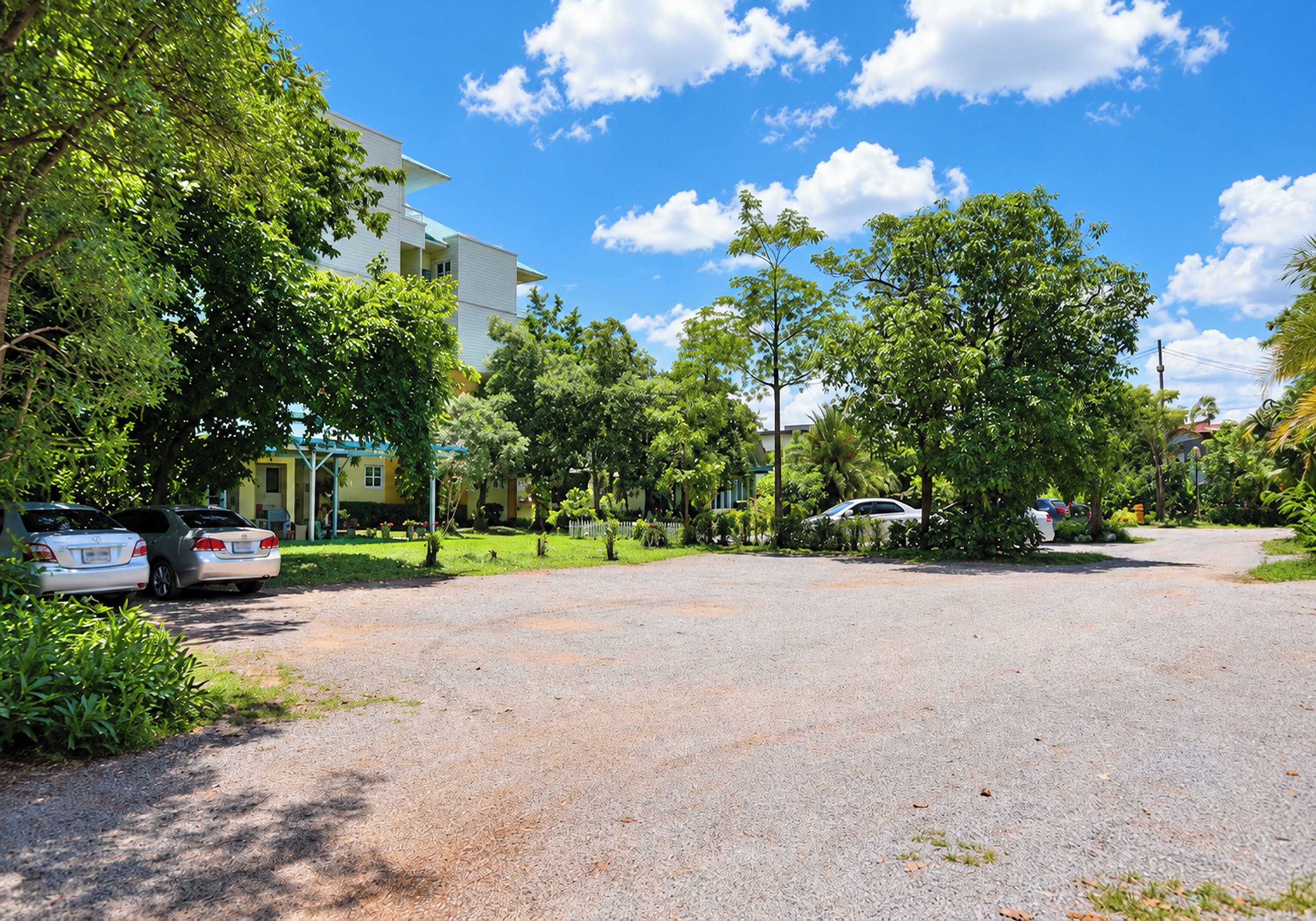 Un camino de entrada residencial con coches estacionados, rodeado de exuberante vegetación y un edificio de varios pisos al fondo.