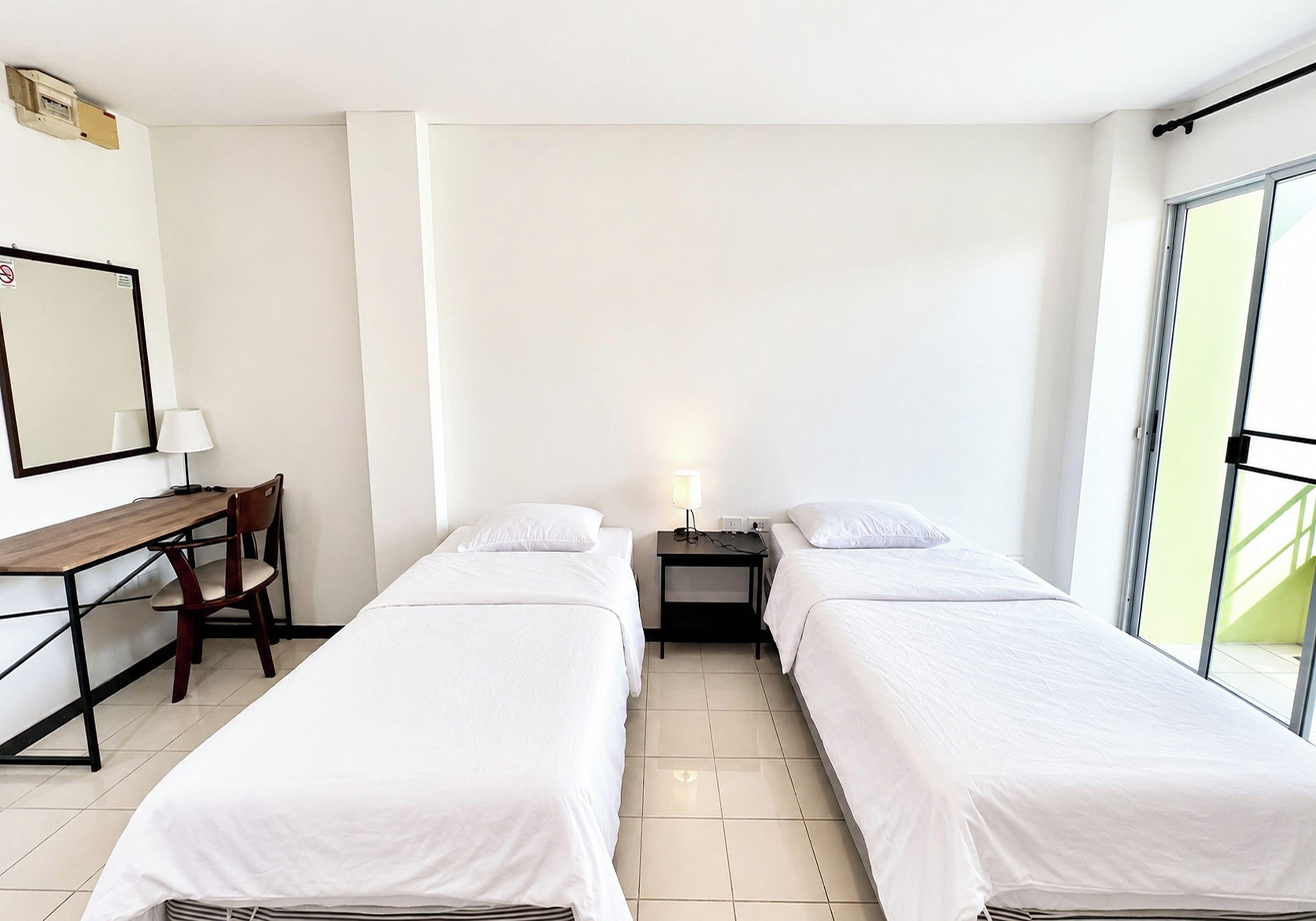 Habitación de hotel con dos camas individuales, un escritorio con silla, una mesita de noche con lámpara y un ventanal grande.