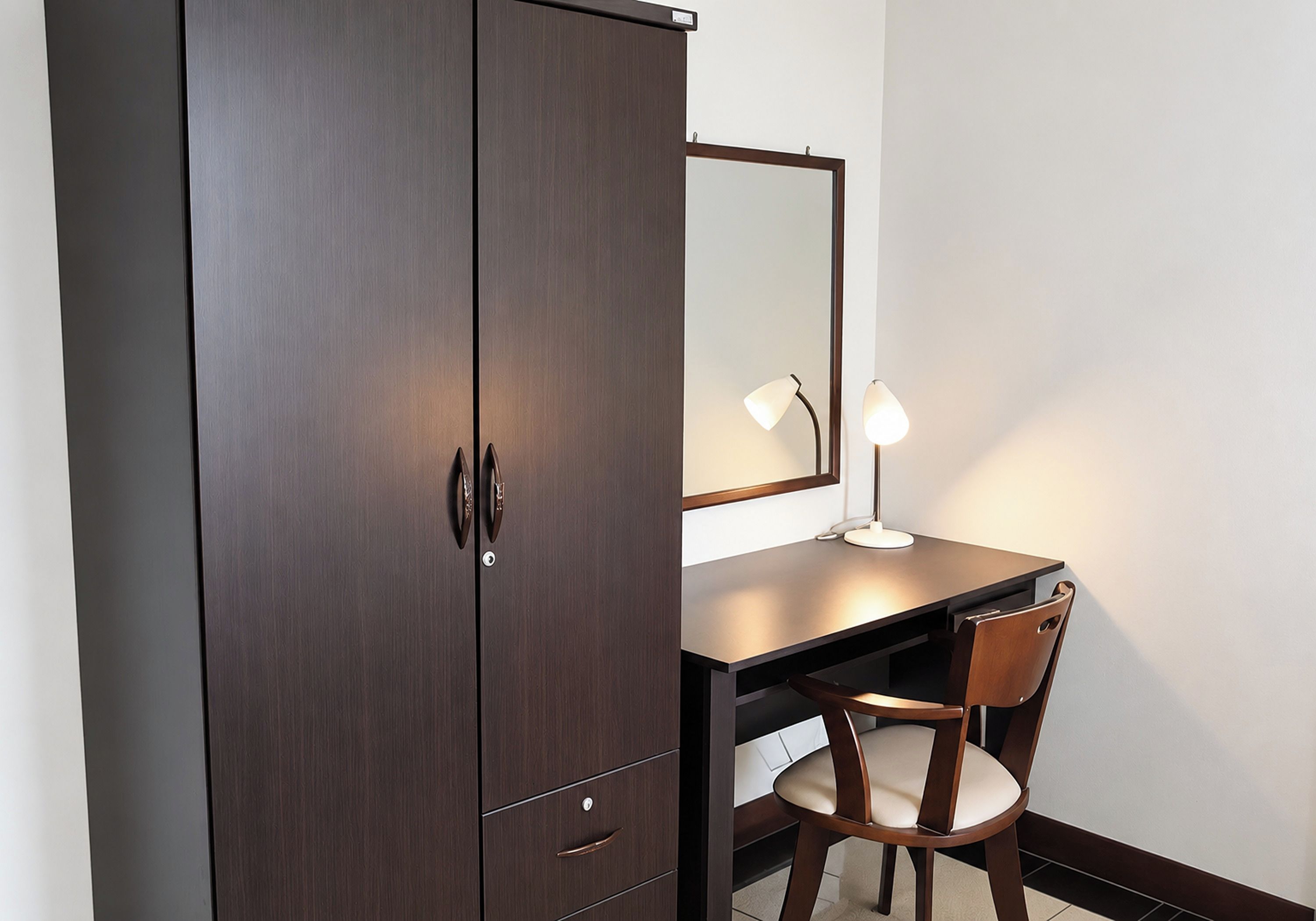 A room with a dark wooden wardrobe, a desk with a chair, and a mirror.
