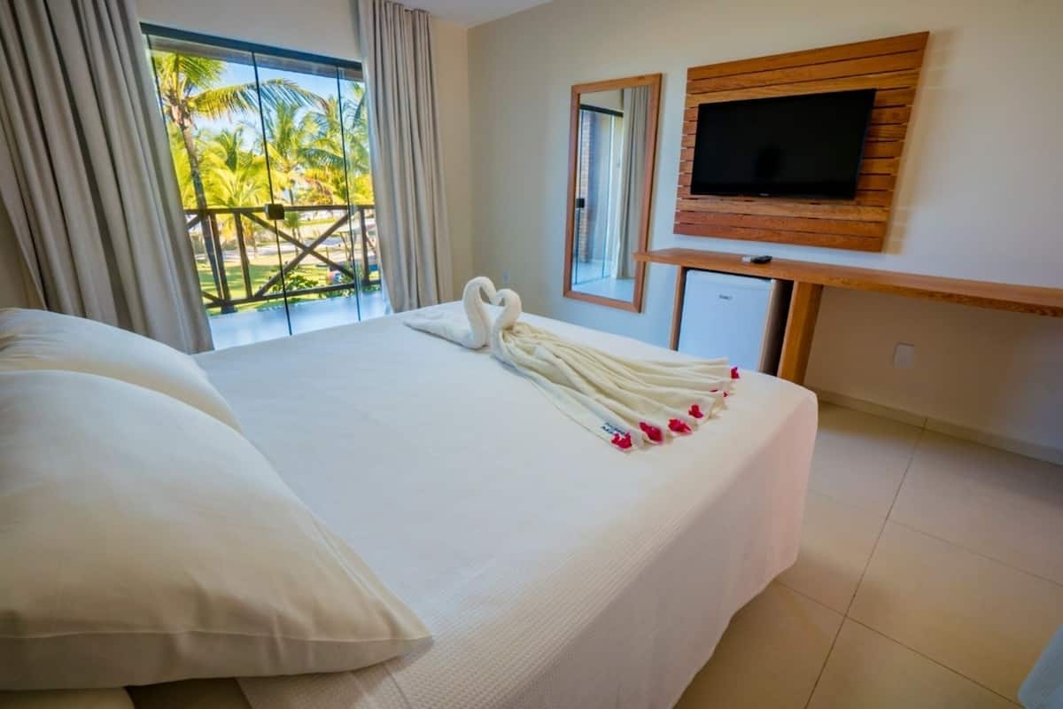 A hotel room with a large bed, a TV mounted on the wall, and a balcony with a view of palm trees.