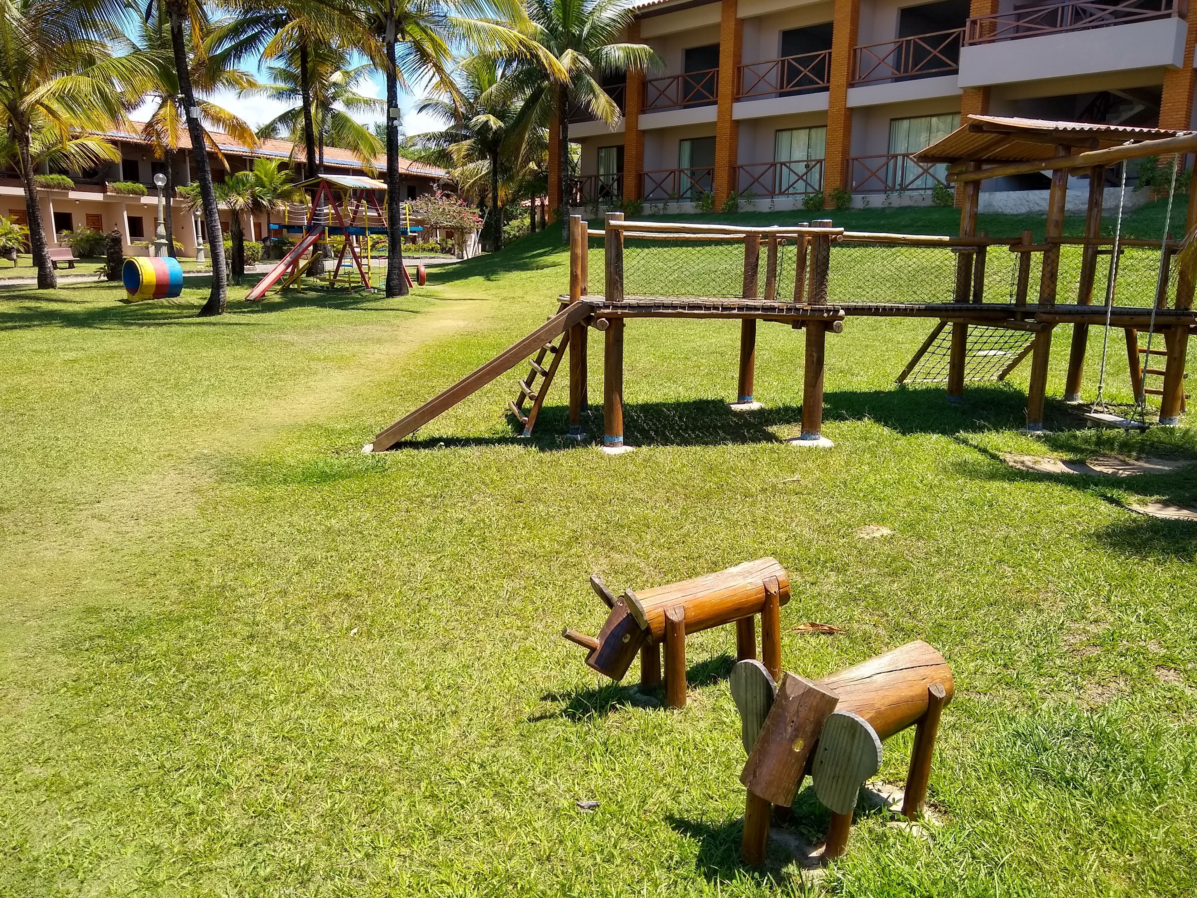 A playground with wooden structures and a fenced area, surrounded by grass and palm trees.