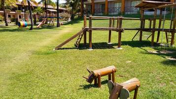 A playground with wooden structures and a fenced area, surrounded by grass and palm trees.