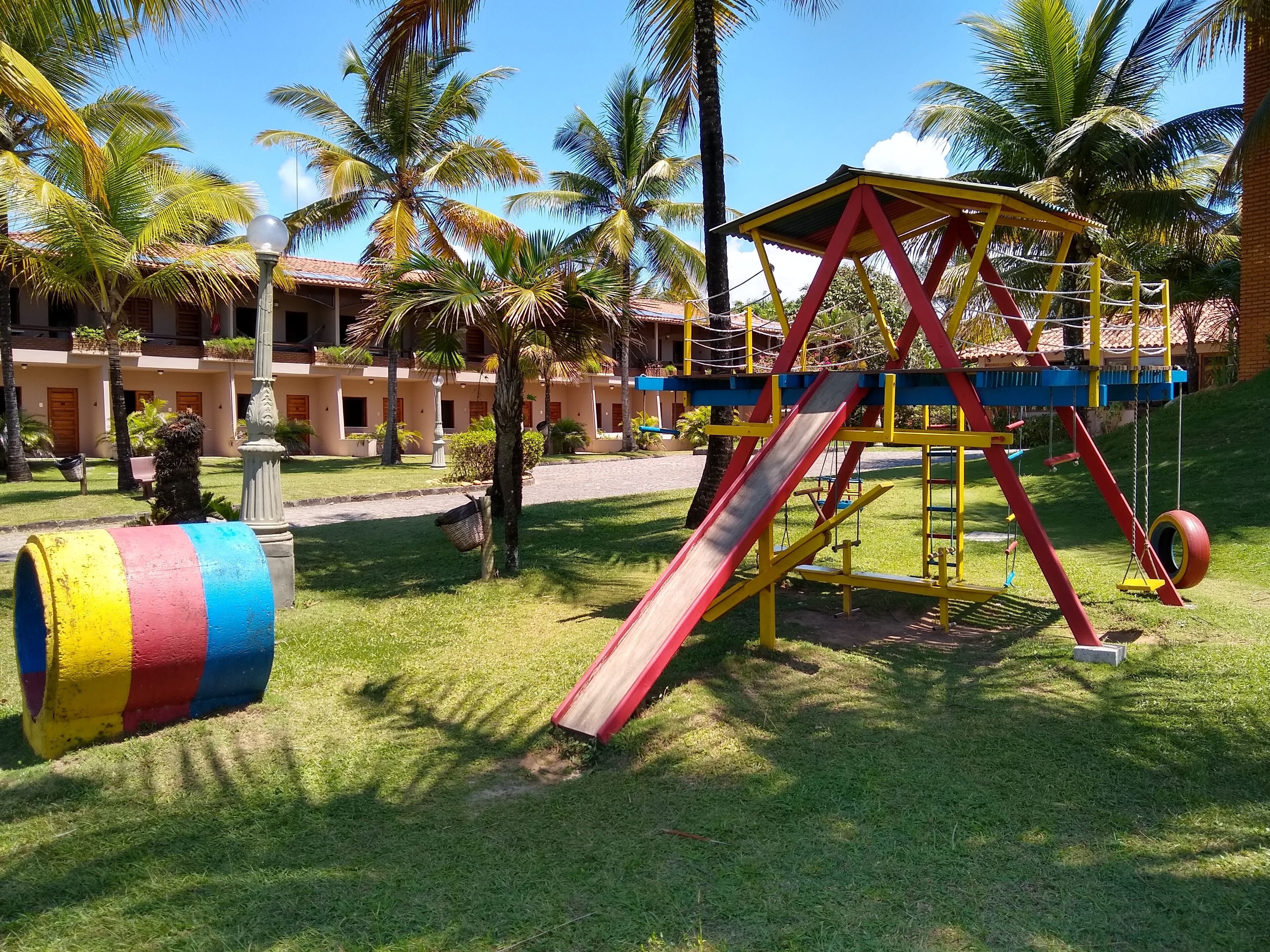 Un parque infantil con tobogán y columpios, rodeado de palmeras y un edificio con techo de tejas.