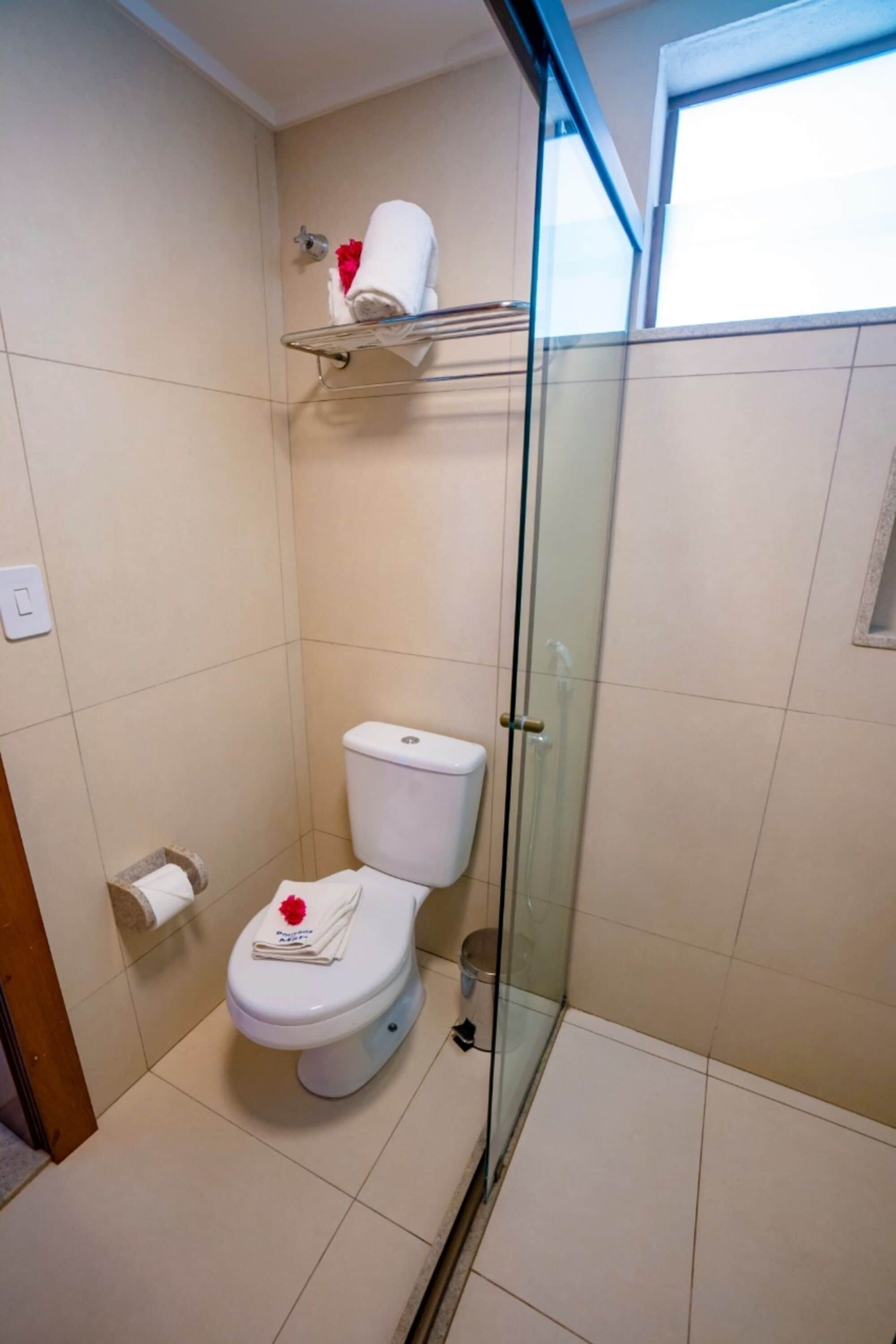 A bathroom with a glass shower enclosure, a toilet with a towel and a red flower on it, and a shelf above the toilet.