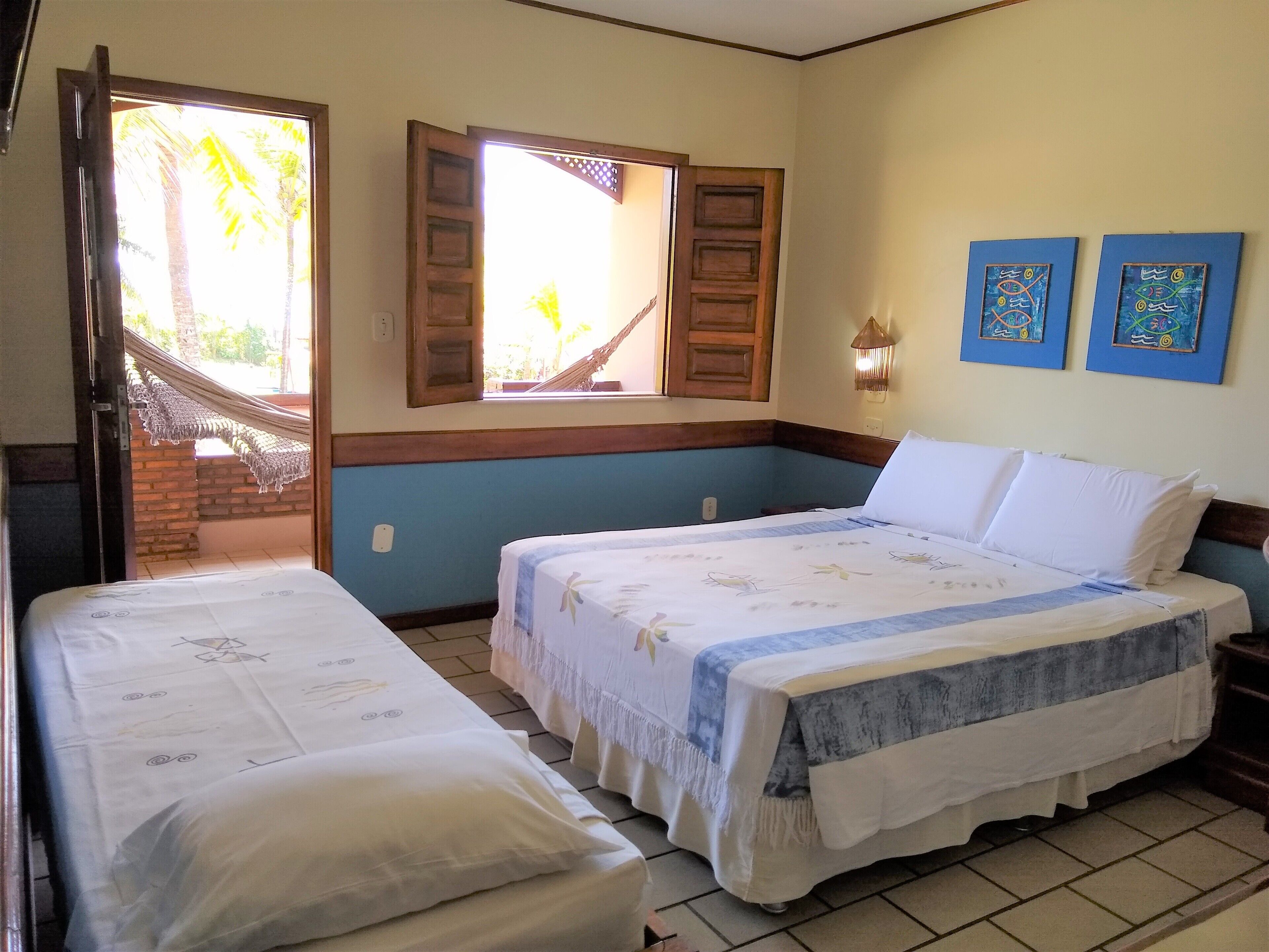 A room with two beds, a window with wooden shutters, a hammock outside, and two wall-mounted artworks.