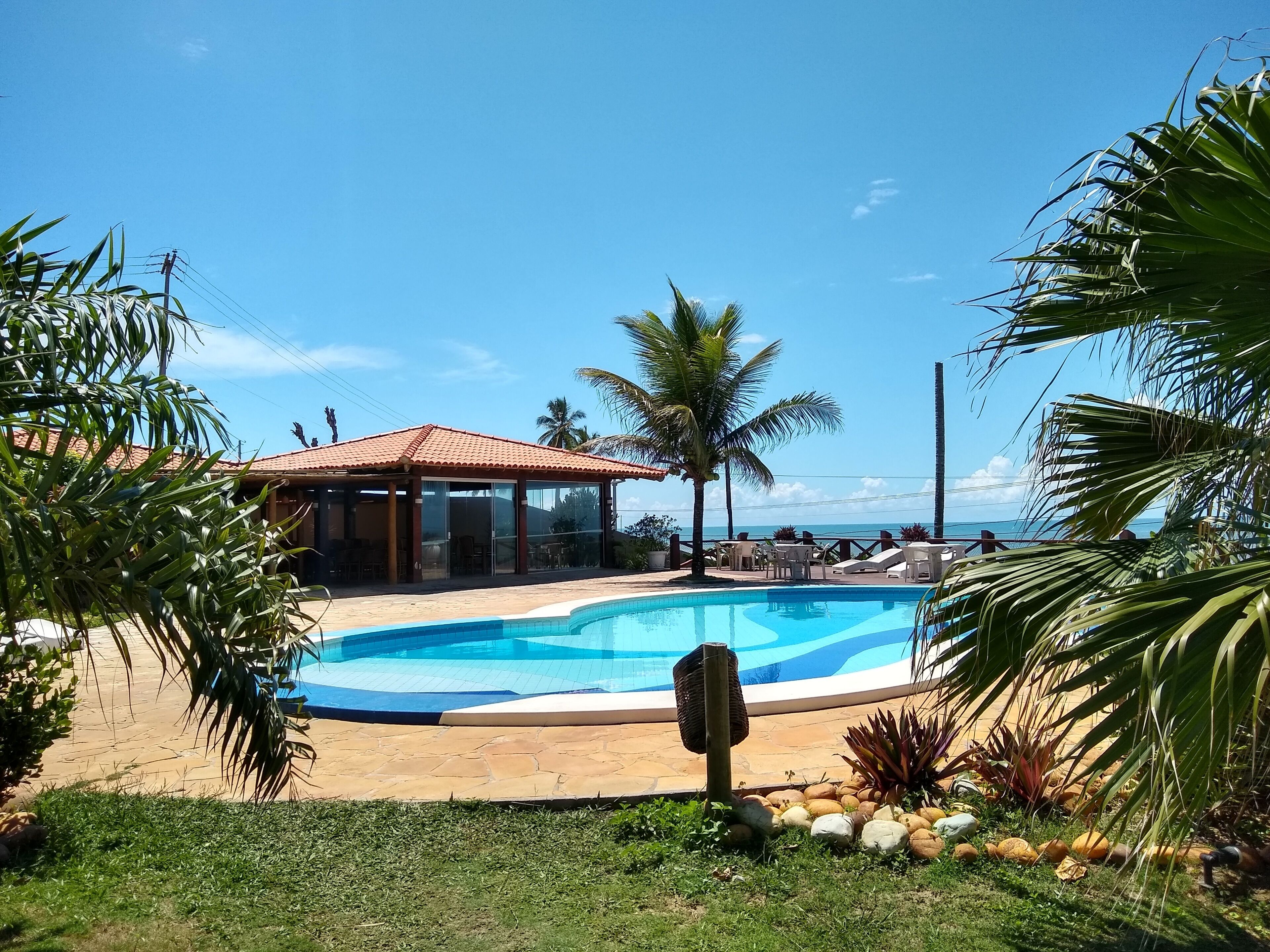 Ein Poolbereich mit überdachter Lounge, Palmen und Blick auf den Ozean.