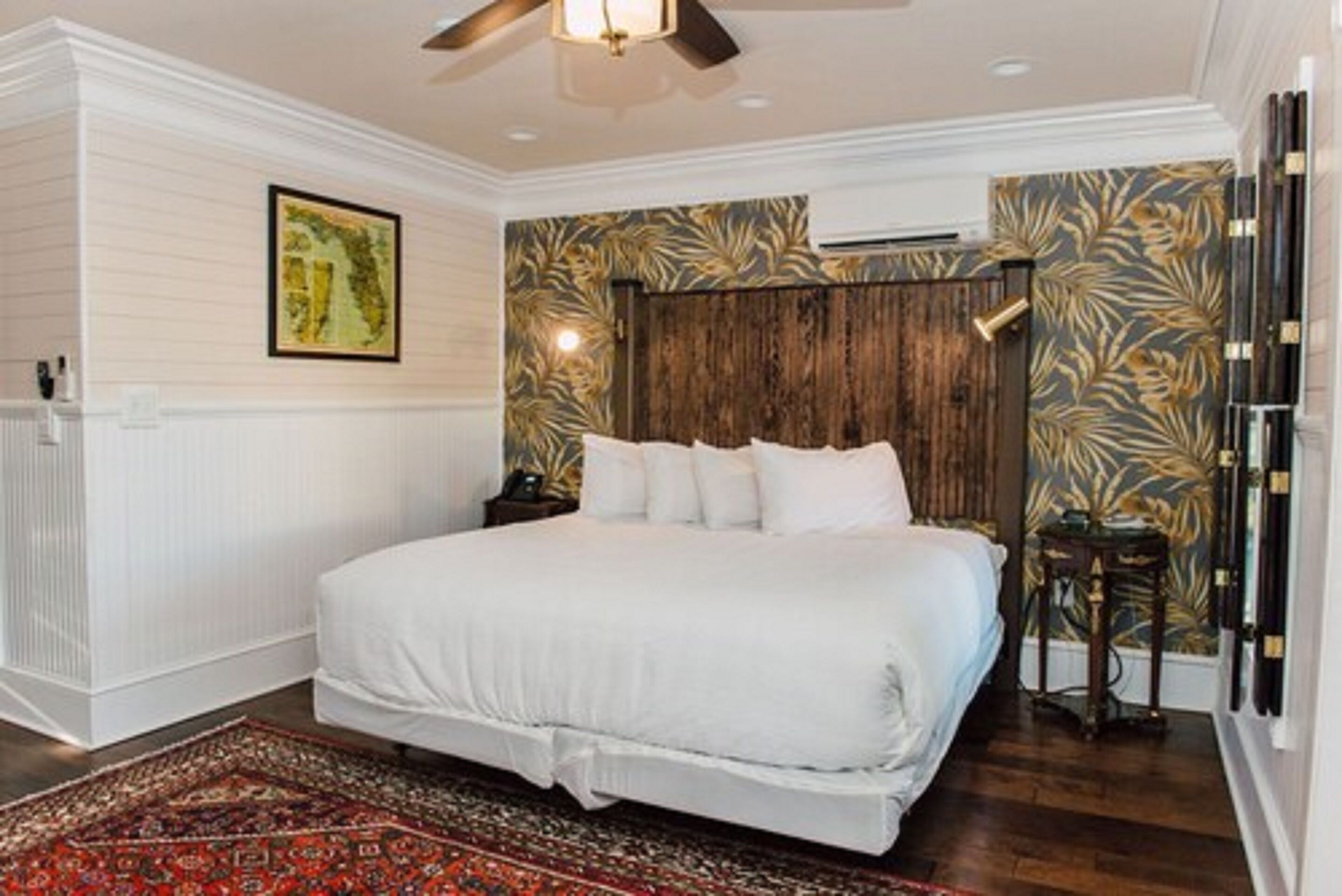 A bedroom with a large bed, a ceiling fan, a nightstand, a wall-mounted air conditioner, and a patterned rug.