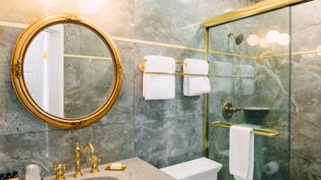 Ein modernes Badezimmer mit Marmorboden, einem goldfarbenen Spiegelrahmen, einer Glasduschkabine und einem Toilettenbecken mit Goldarmatur.