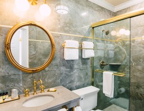 Moderni kylpyhuone, jossa on marmorilattia, kultakehyksinen peili, lasiseinäinen suihku ja kultainen hana varustettu wc-istuin.