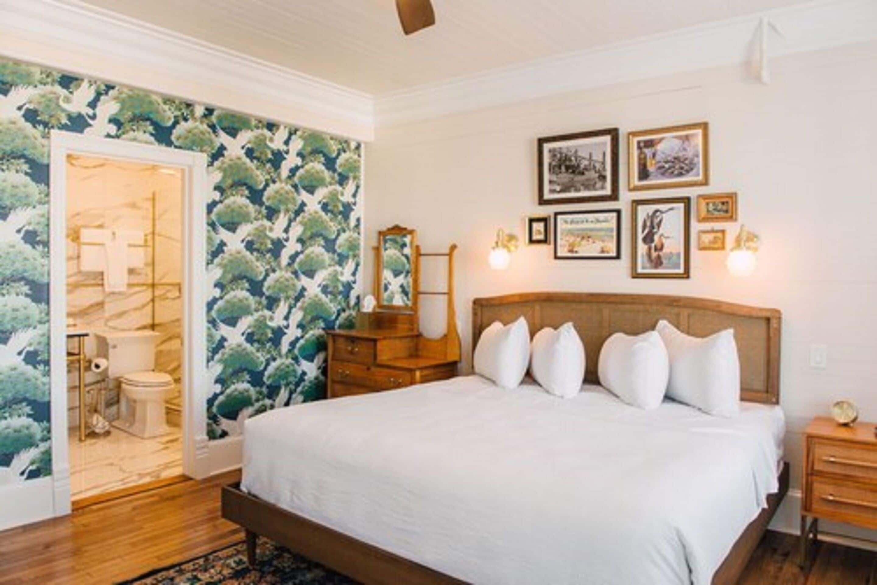 A bedroom with a wooden bed, white bedding, a wooden dresser, and a mirror. There are framed pictures on the wall and a door leading to a bathroom.