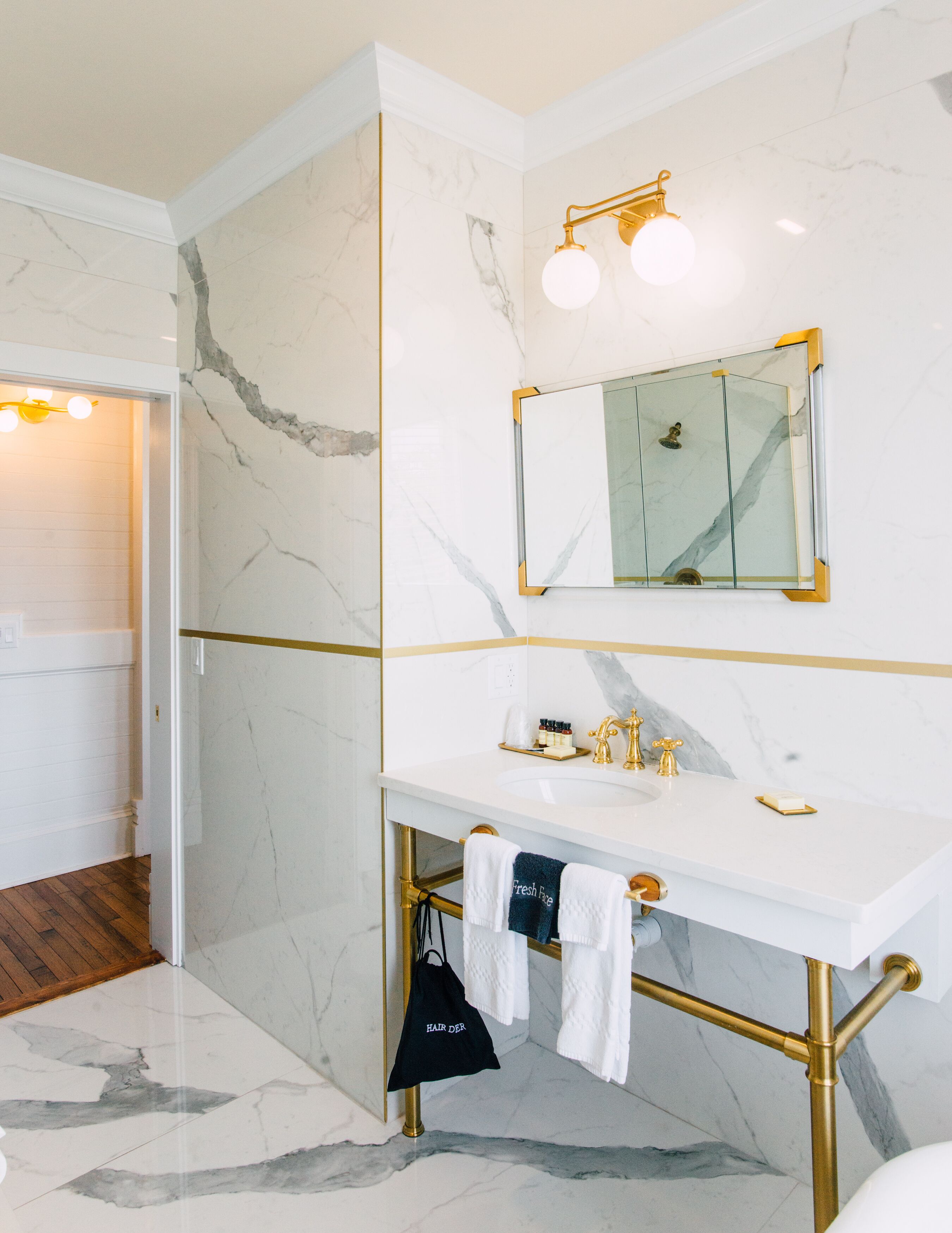 Une salle de bain avec un mur en marbre blanc, des accessoires dorés et un grand miroir.