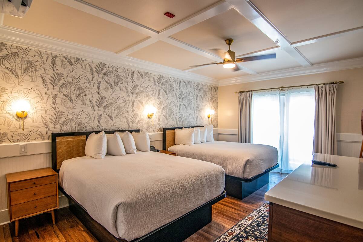 A hotel room with two beds, a desk, a ceiling fan, and a patterned wallpaper.