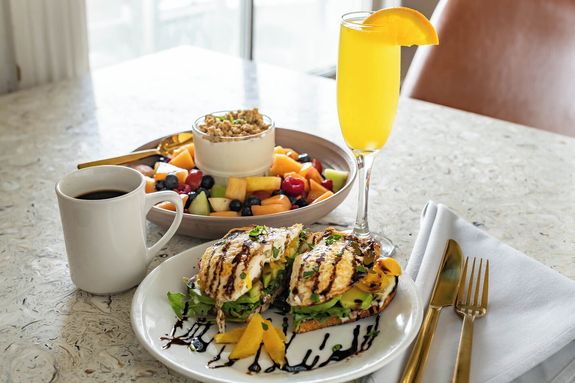 A breakfast setting with a plate of grilled chicken sandwich, a bowl of fruit, a glass of orange juice, and a cup of coffee.