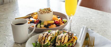 A breakfast setting with a plate of grilled chicken sandwich, a bowl of fruit, a glass of orange juice, and a cup of coffee.