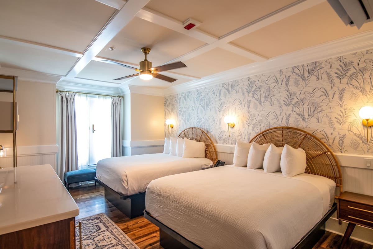 A hotel room with two beds, a ceiling fan, a large window with curtains, and a wall with a floral pattern.