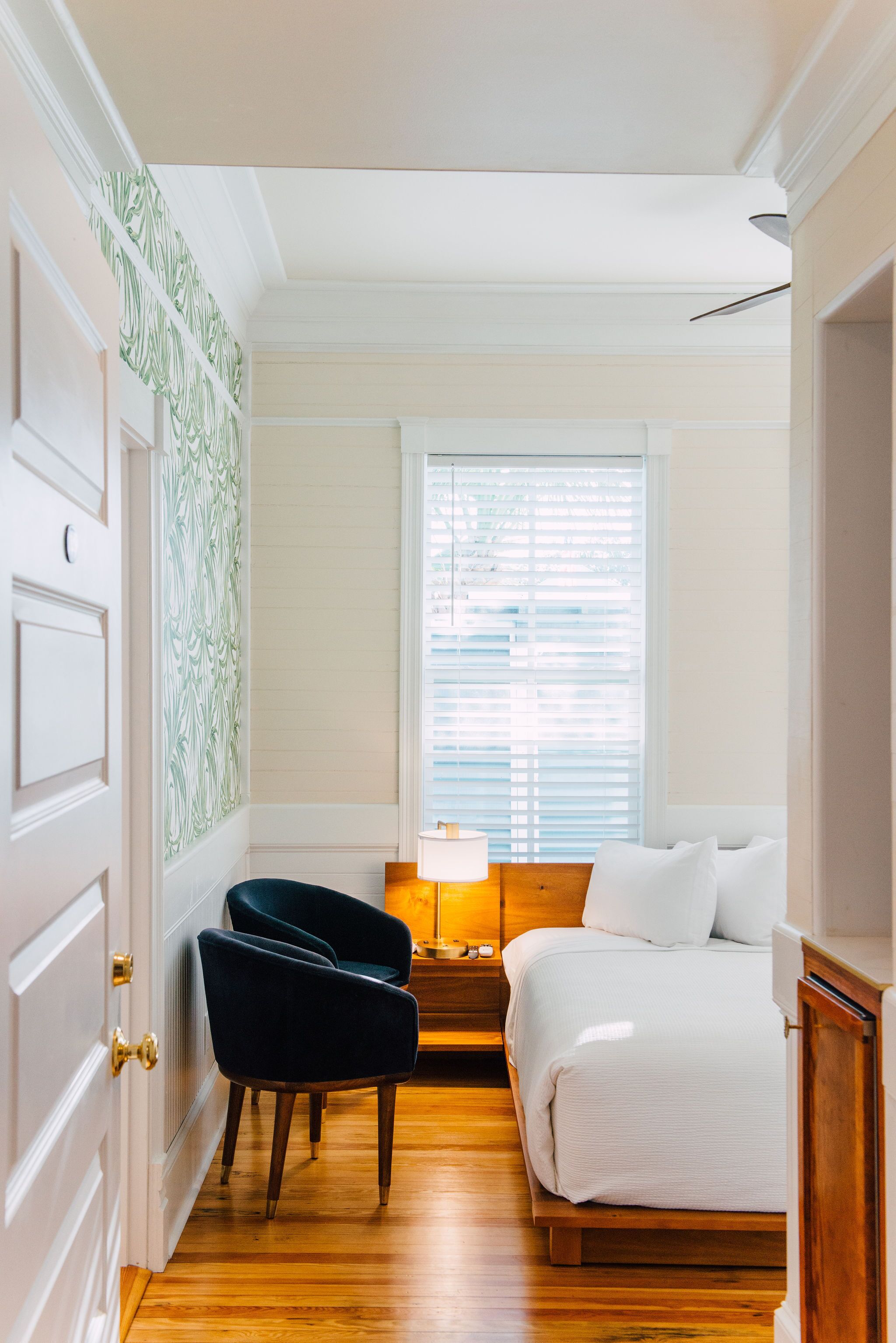 A bedroom with a bed, two chairs, a nightstand, a lamp, and a window with blinds.