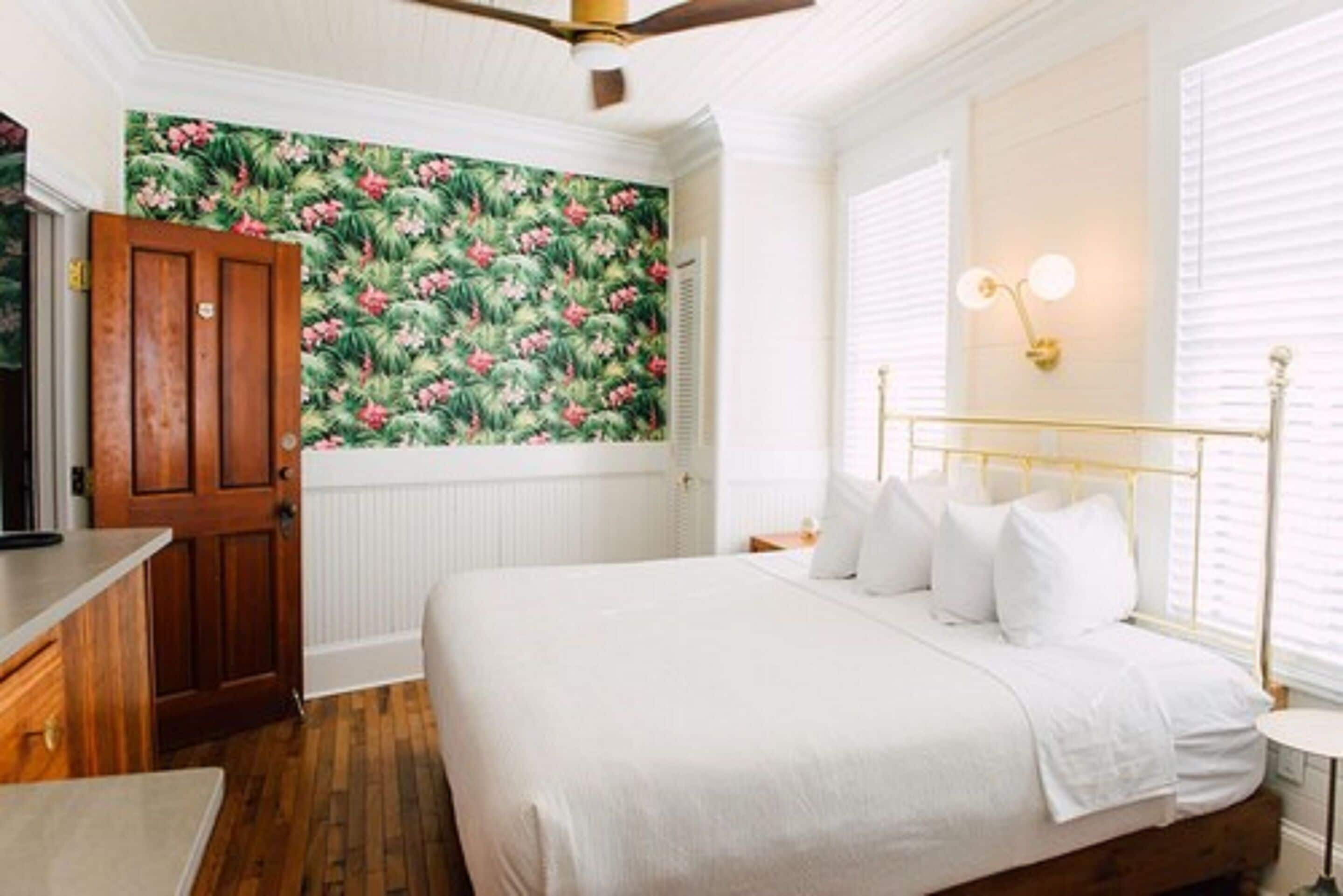 Une chambre avec une porte en bois, un lit avec une literie blanche, un ventilateur de plafond et un mur recouvert d’un papier peint à motifs floraux.