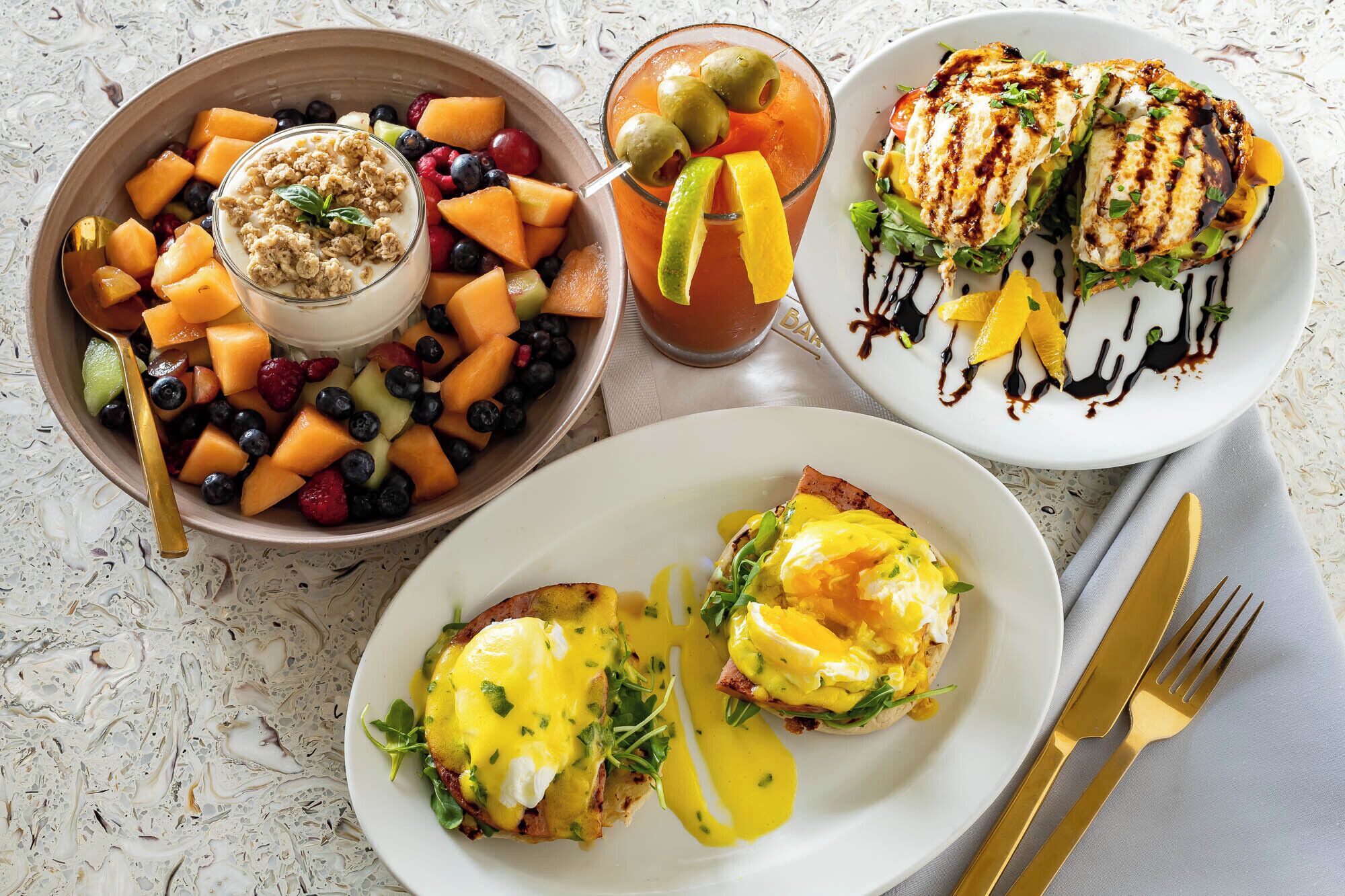 Une table mise pour le petit-déjeuner avec un saladier de salade de fruits, un verre de jus de fruits, ainsi que des assiettes contenant des œufs Bénédicte et du poulet grillé.
