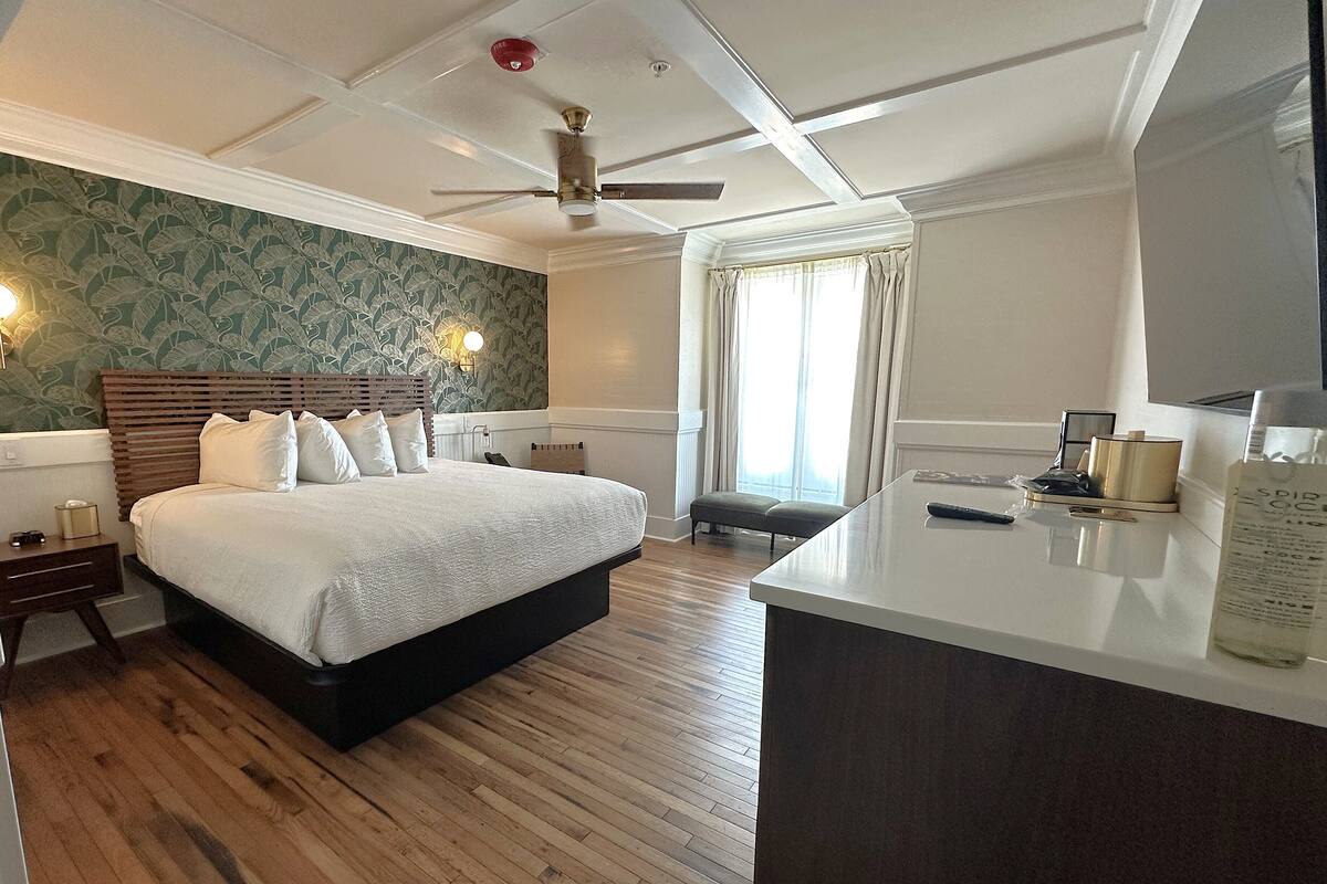 A hotel room with a large bed, wooden flooring, a ceiling fan, a desk with a lamp, and a window with curtains.