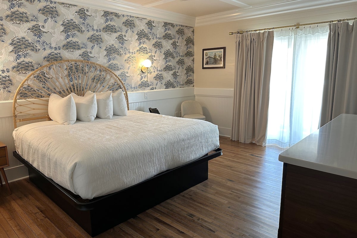 A bedroom with a large bed, a bedside table, a chair, a desk, a ceiling fan, and a window with curtains.