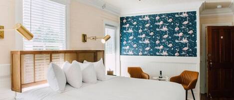 Une chambre avec un grand lit, un ventilateur de plafond, une fenêtre avec des stores, un mur recouvert d’un papier peint à motifs floraux et un plancher en bois.
