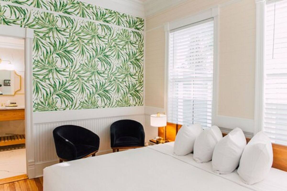 A hotel room with a bed, two black chairs, a nightstand, a lamp, and a window with blinds.